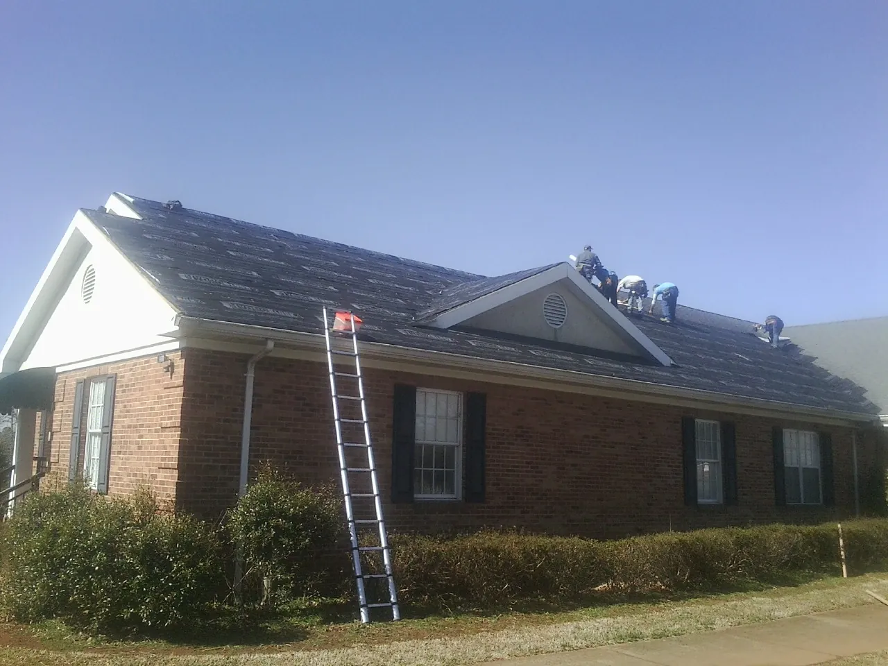 Crew tearing shingles off a brick ranch roof with a ladder set — Metal Roof Repair work by our crew in Colebrook
