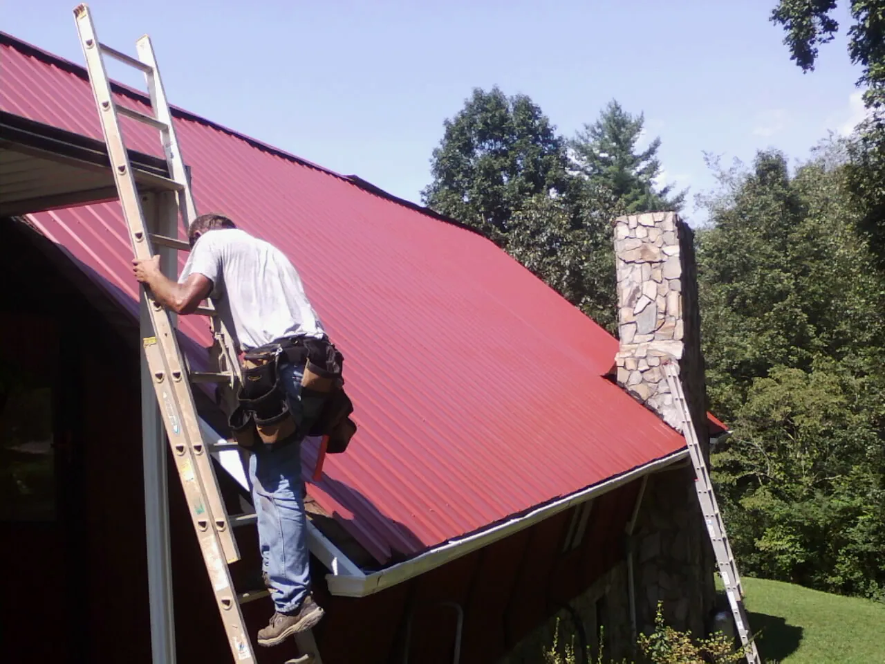 Roofer on a ladder beside a red metal roof and stone chimney — Soffit & Fascia Repair by Roofing Brothers in Colebrook